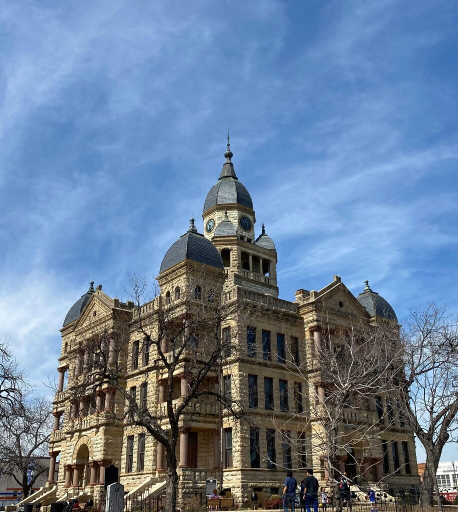 Low angle view of Denton County Courthouse against blue sky, featuring bare trees in winter.