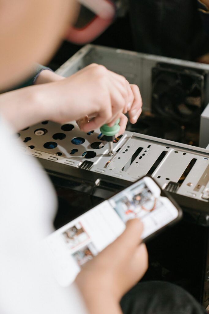 Hands using screwdriver to repair computer hardware, referencing smartphone for guidance.